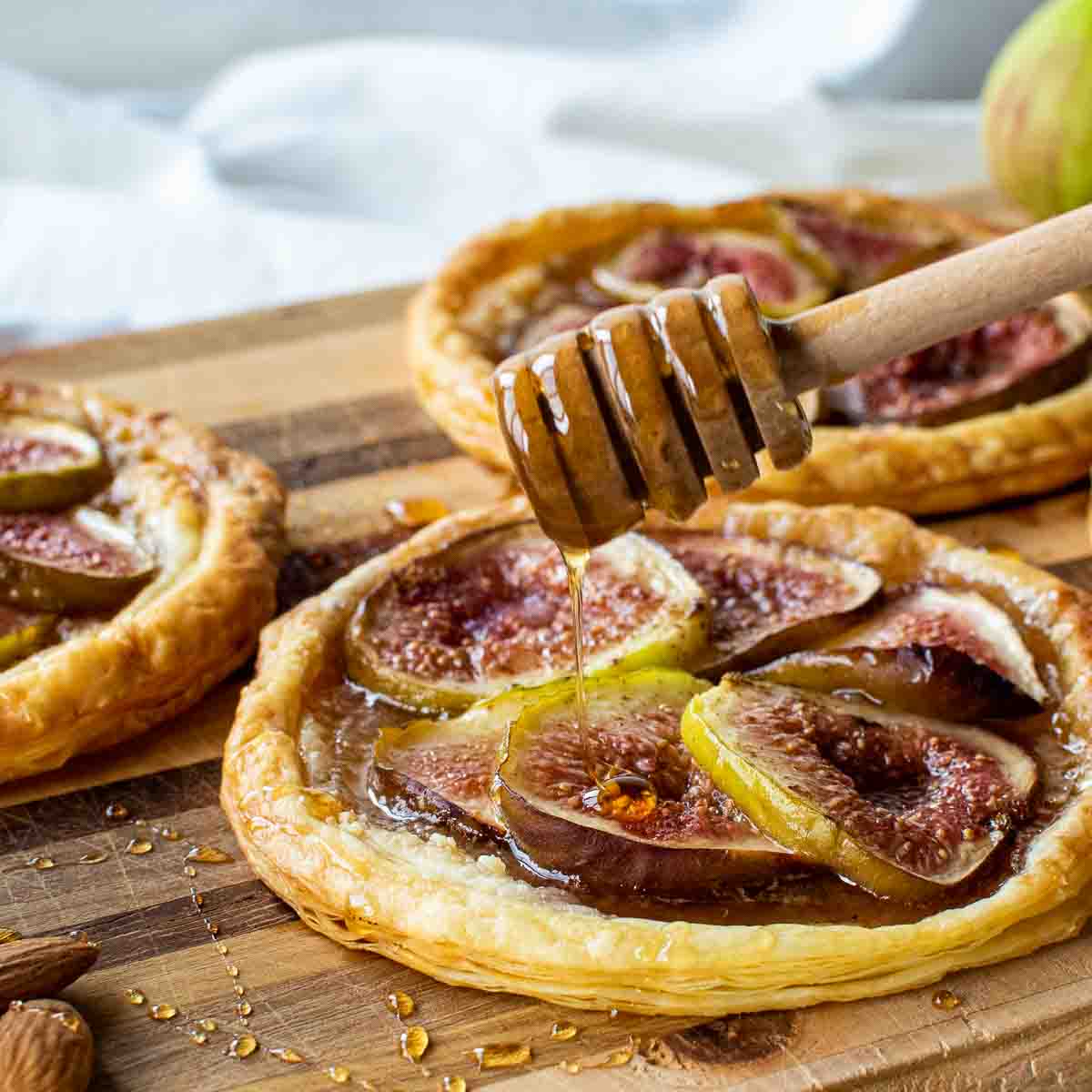 puff pastry fig tartlets on a wooden board with honey dipper drizzling honey over pastries.