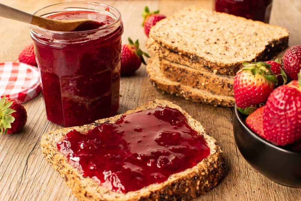How to make microwave strawberry jam using just 3 ingredients #strawberryjam #microwavejam #jam #strawberryrecipes Bread served with the preserves and a jar on a wooden table.