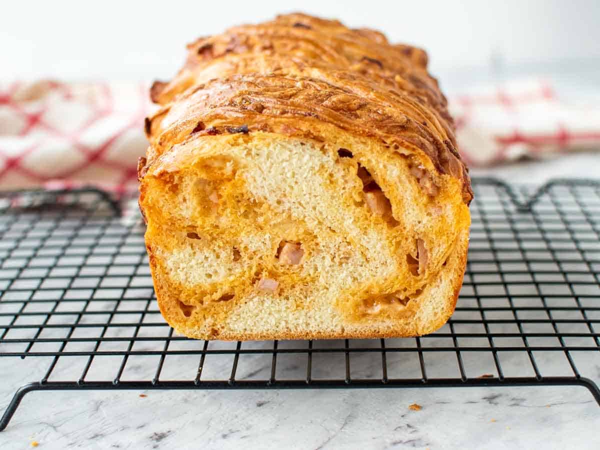 This savoury Cheese Bread with Bacon is beautiful to look at and super tasty to eat. The dough is enriched with butter and egg then rolled up with cheese and bacon. You won't be able to stop eating this one! #bread #cheesebread #bakingwithyeast #bake Cross section of cheese filled Bread loaf on black wire rack.