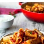 Rigatoni Arrabbiata on a white plate with red skillet in the background.
