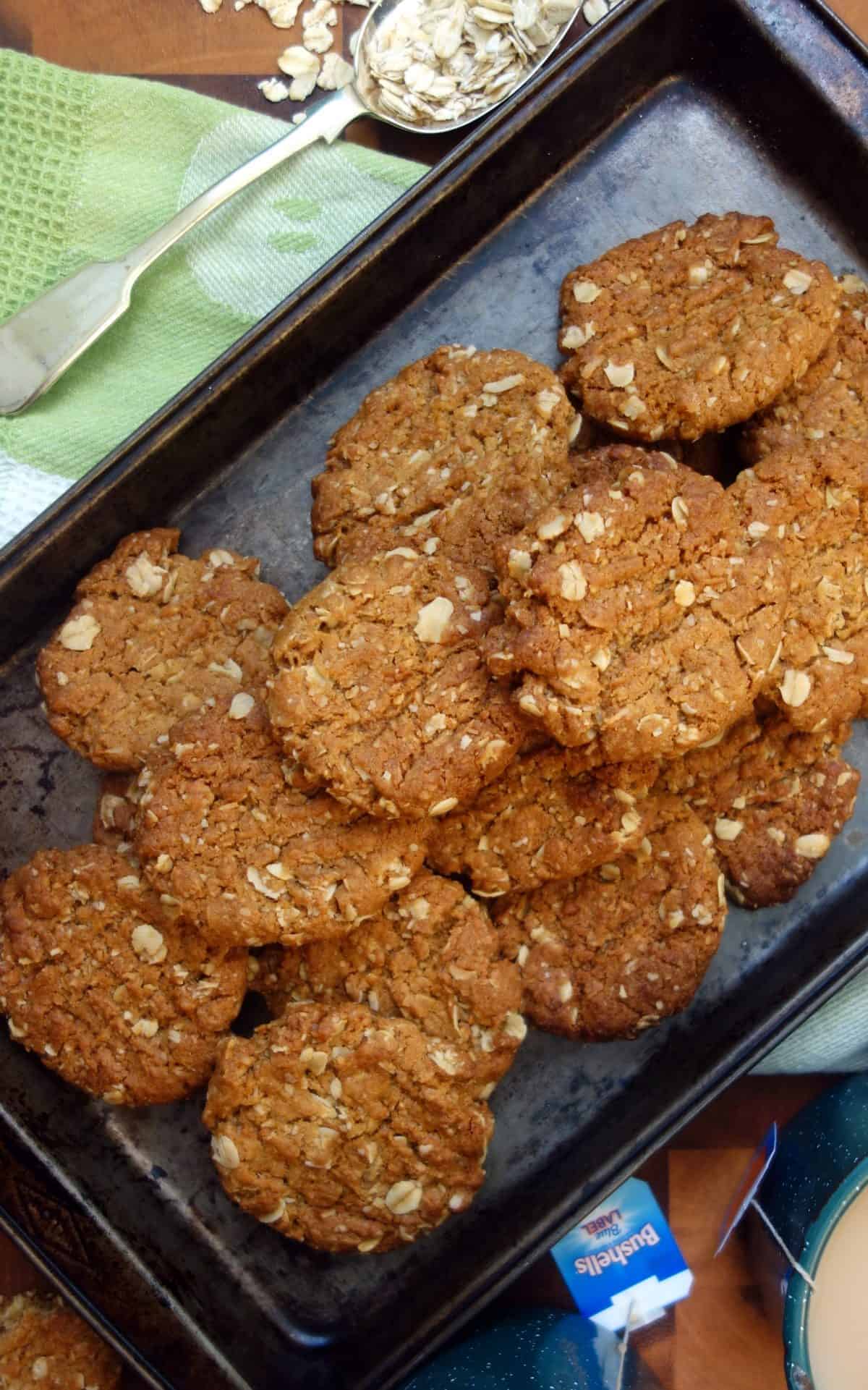 These Anzac Biscuits are beautifully flavored with golden syrup and ground ginger. #anzacbiscuits #cookies #biscuits #recipe #australianrecipe Overhead view of Anzac Biscuits in dark coloured baking tray
