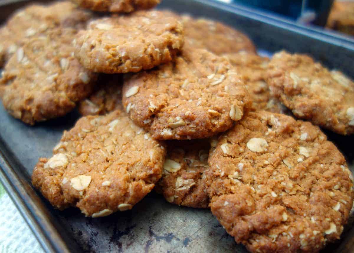 These Anzac Biscuits are beautifully flavored with golden syrup and ground ginger. #anzacbiscuits #cookies #biscuits #recipe #australianrecipe Close up of Anzac Biscuits on dark coloured baking tray