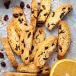 Orange Cranberry Biscotti on white paper with halved oranges and dried cranberries scattered viewed from above.