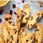 Orange Cranberry Biscotti on white paper with halved oranges and dried cranberries scattered viewed from above.