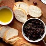white bowl of olivada, bread, oil, parsley and olives all on a wooden board
