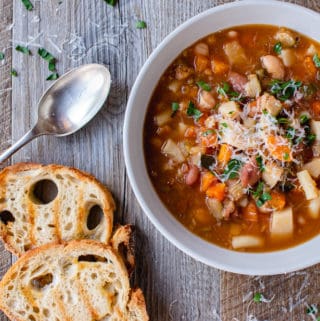 White bowl of slow cooker minestrone soup, two slices of toasted bread and spoon on wooden table