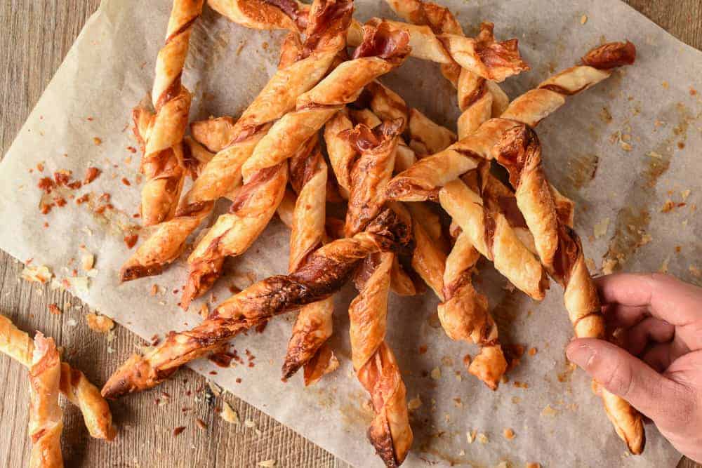Crunchy Puff Pastry Twists with Prosciutto and Parmesan are delicious with drinks or as a snack. Plus so easy to make! #recipe #puffpastry #prosciutto #appetizer #snack #cheese pile of pastry sticks with hand reaching in to take one.