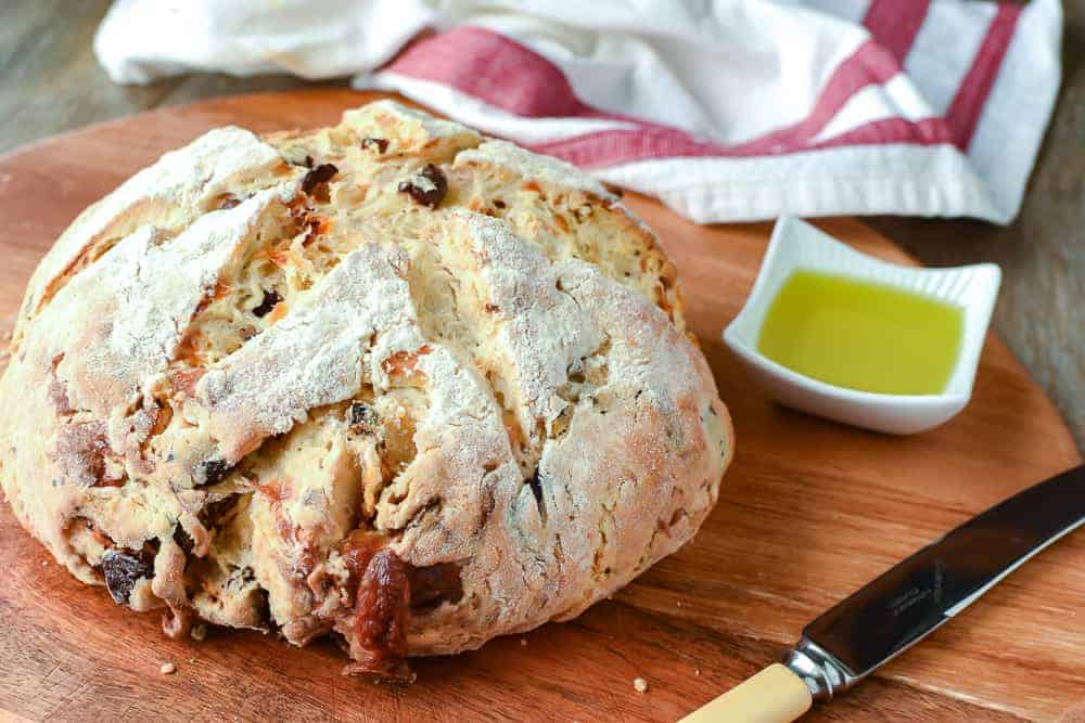 Damper with olives, sun dried tomatoes and cheese is tasty, quick and delicious #damper #quickbread #sodabread #recipe #italianfood #australianfood Damper bread on wooden board with knife, white dish of olive oil, dish clot in the background.