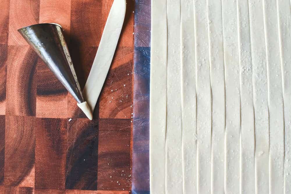 These Mini Cream Horns use store bought puff pastry taking out all the difficulties of pastry. Fill with fresh cream or my special buttercream for a delicious treat. #creamhorns #minicreamhorns #puffpastryrecipes #puffpastry #recipe Sheet of raw puff pastry cut into strips with one strip being wound around a mould viewed from above.