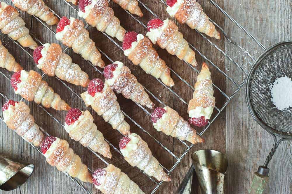 These Mini Cream Horns use store bought puff pastry taking out all the difficulties of pastry. Fill with fresh cream or my special buttercream for a delicious treat. #creamhorns #minicreamhorns #puffpastryrecipes #puffpastry #recipe Mini cream horns filled with cream and topped with raspberry on a wire rack viewed from above.
