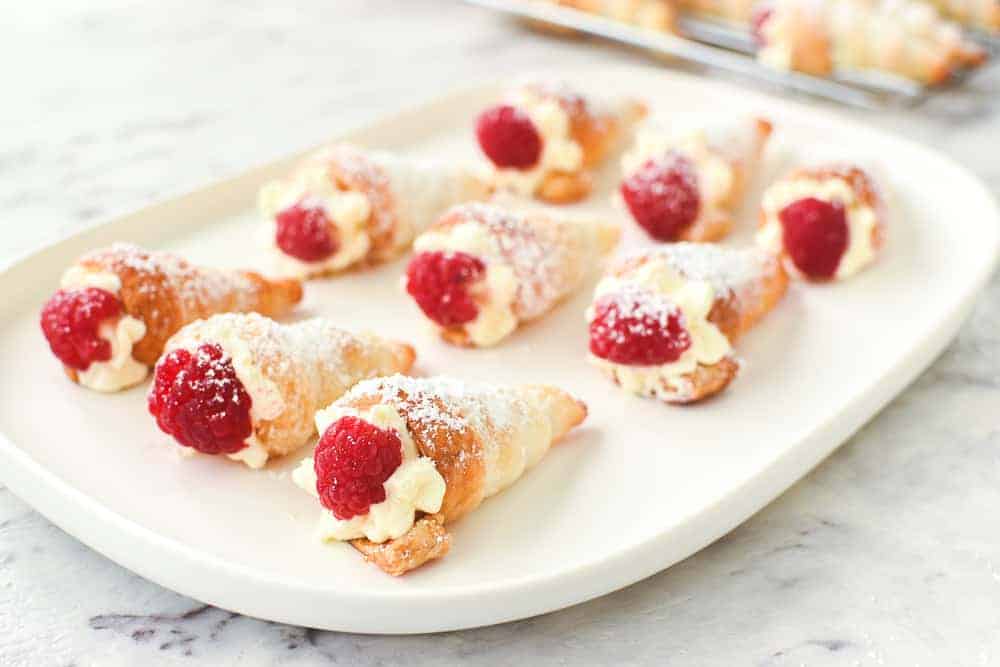 These Mini Cream Horns use store bought puff pastry taking out all the difficulties of pastry. Fill with fresh cream or my special buttercream for a delicious treat. #creamhorns #minicreamhorns #puffpastryrecipes #puffpastry #recipe Puff pastry cones filled with cream and topped with raspberry on an oblong white plate viewed from an angle.