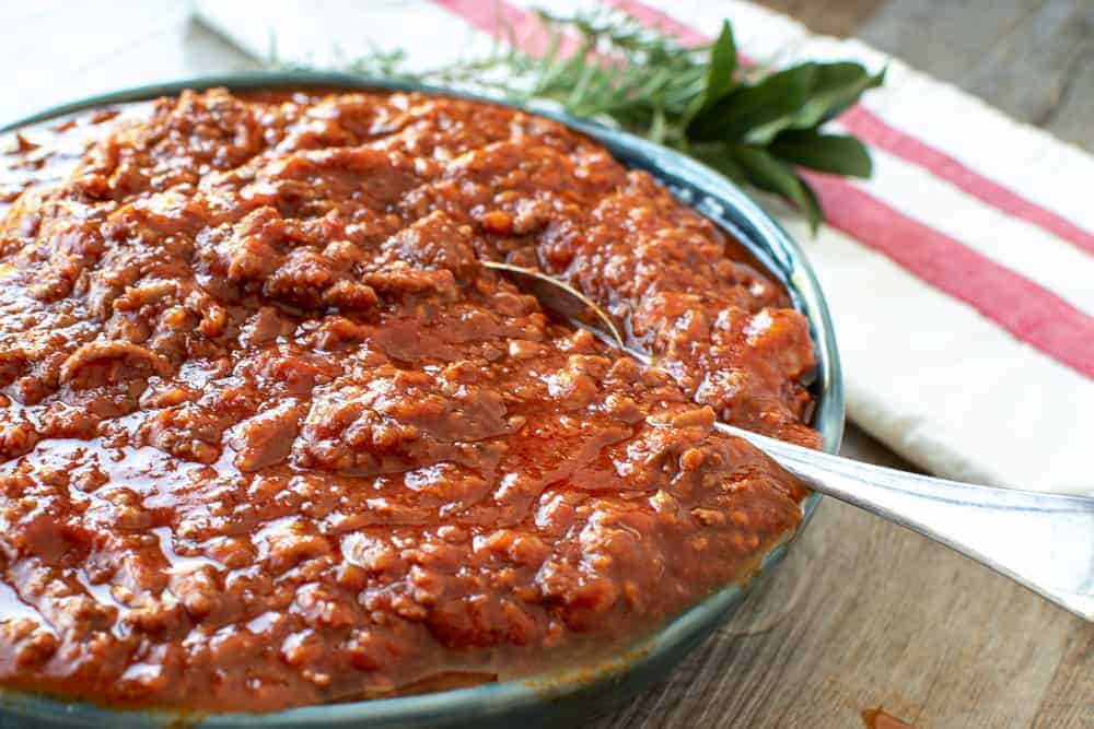 This Ragù Pasta Sauce has my father's secret ingredient that makes all the difference. This recipe makes a big batch so that you have lots to freeze for later. #ragu #pastasauce #italian #recipe blue bowl overflowing with bolognese sauce, spoon in the sauce and herbs in the background.