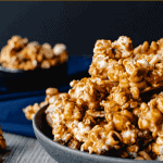 Homemade caramel popcorn in a grey bowl on blue cloth. smaller black bowl of caramel popcorn in background caramel popcorn and nuts on wooden table. close up