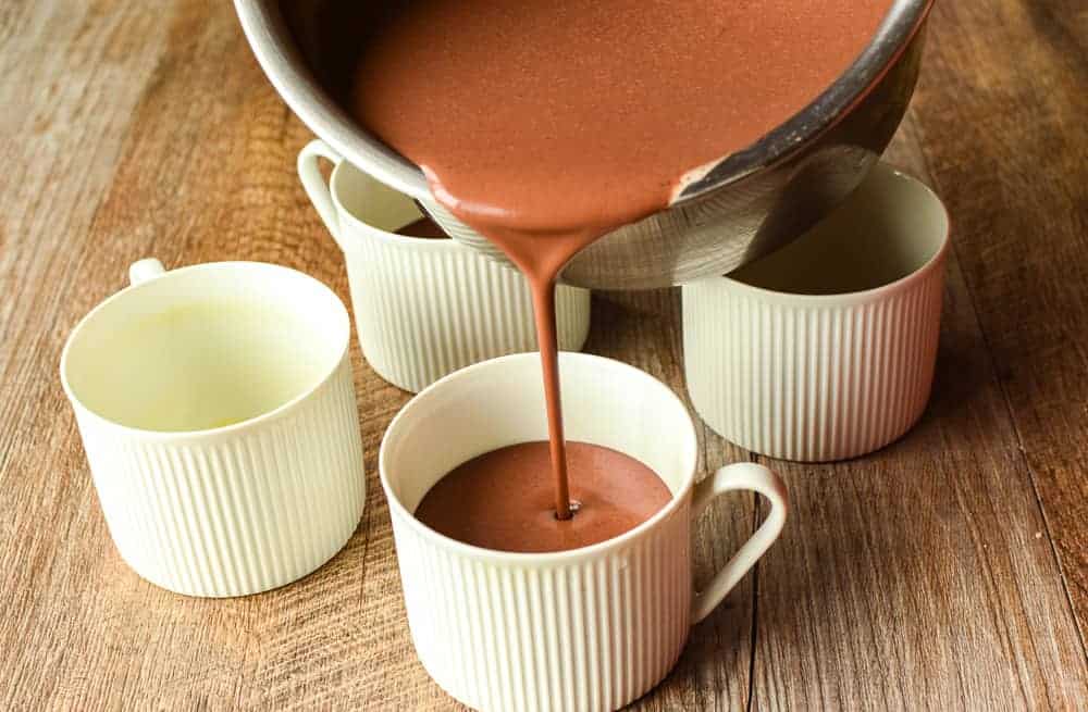 Budino al Cioccolato is a rich, smooth chocolate baked custardloved by children and adults alike. #budinoalcioccolato #chocolatepudding #pudding #recipe #bakedcustard four white teacups on wooden table and chocolate being poured into one of the teacups from a stainless steel saucepan