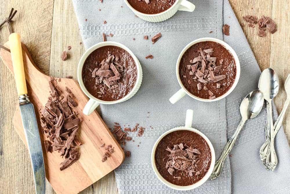 Budino al Cioccolato is a rich, smooth chocolate baked custardloved by children and adults alike. #budinoalcioccolato #chocolatepudding #pudding #recipe #bakedcustard four teacups filled with chocolate pudding on a grey cloth with a wooden board of chopped chocolate viewed from above