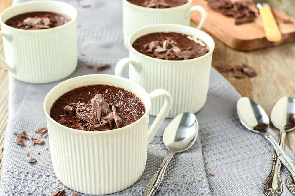 Budino al Cioccolato is a rich, smooth chocolate baked custardloved by children and adults alike. #budinoalcioccolato #chocolatepudding #pudding #recipe #bakedcustard four teacups filled with chocolate pudding on a grey cloth with a wooden board of chopped chocolate and four silver spoons viewed from front on