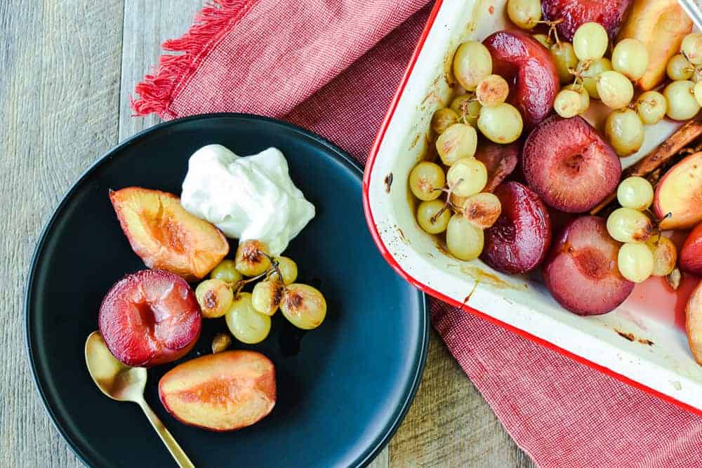 Roasted Fruit with Prosecco and Honey is an easy dessert with vanilla ice cream and leftovers are particularly good for breakfast the next day. #fruit #summerfruit #stonefruit #grapes #roastedfruit #roasted #dessert #breakfast roasted plums, peaches and green grapes with scoop of yogurt on dark plate with gold spoon, on the right is a white pan with red trim filled with roasted plums, peaches and green grapes on red cloth. All is on a timber table and viewed from above