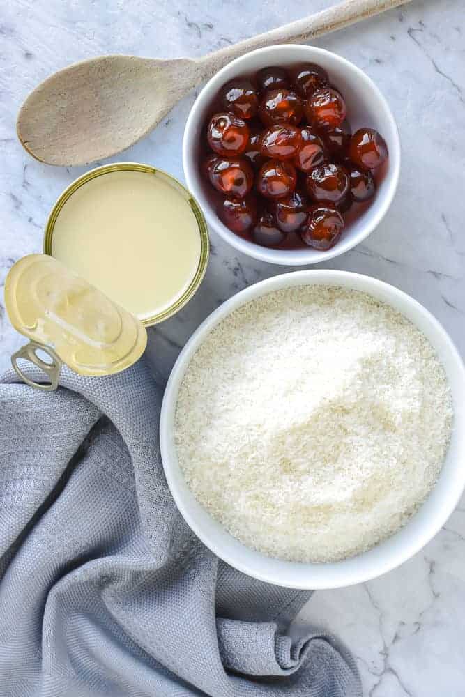 Chocolate Cherry Slice is a wonderful combination of the classic flavors of chocolate, cherry and coconut in one sweet slice. #chocolateslice #chocolatebars #chocolatecherryslice #chocolaterecipe #recipe #baking white bowl of coconut, white bowl of candied cherries, opened can of condensed milk, wooden spoon and grey cloth viewed from above