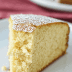 half eaten slice of plain cake on white plate with fork on red cloth with whole cake in the background