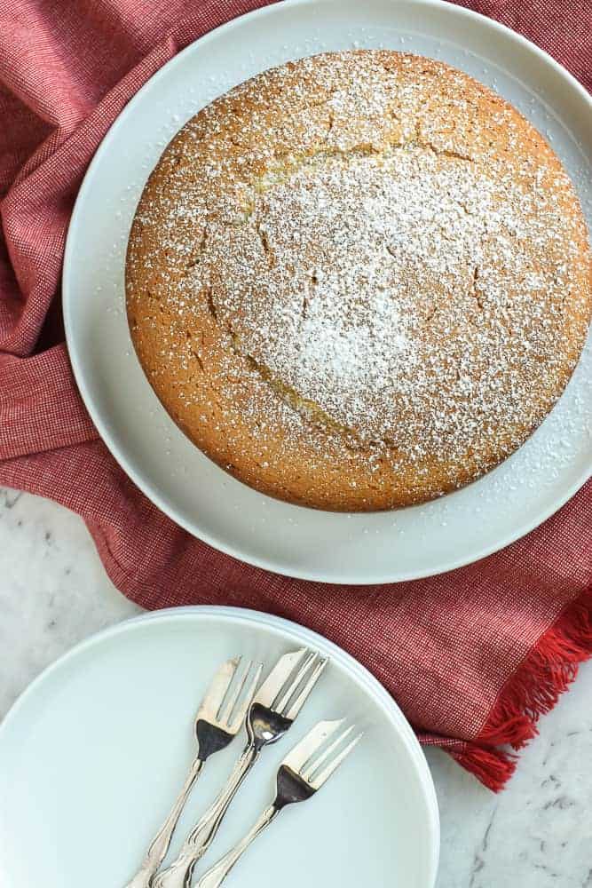 This Easy Vanilla Cake recipe is a classic and comes together in minutes. Even if you've never baked a cake from scratch, you will find this simple and achievable! #easyvanillacake #vanillacake #easycakerecipe #easyrecipe round vanilla cake dusted with powdered sugar on white plate on red cloth with white small plate and forks on the side viewed from above.