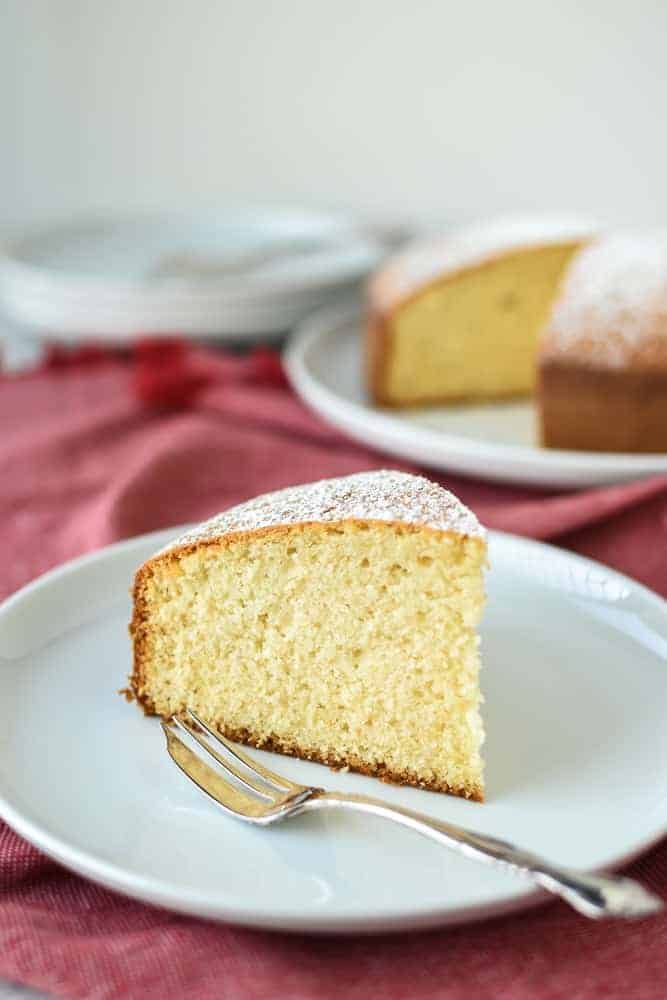 slice of plain cake on white plate with fork on red cloth with whole cake and extra plates in the background