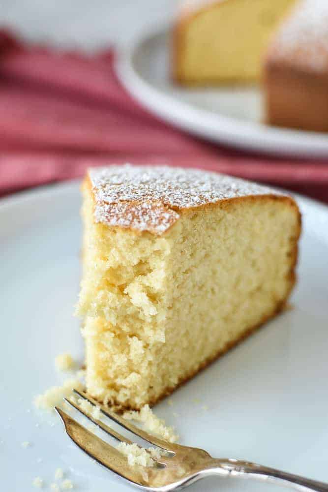 This Easy Vanilla Cake recipe is a classic and comes together in minutes. Even if you've never baked a cake from scratch, you will find this simple and achievable! #easyvanillacake #vanillacake #easycakerecipe #easyrecipe half eaten slice of plain cake on white plate with fork on red cloth with whole cake in the background.