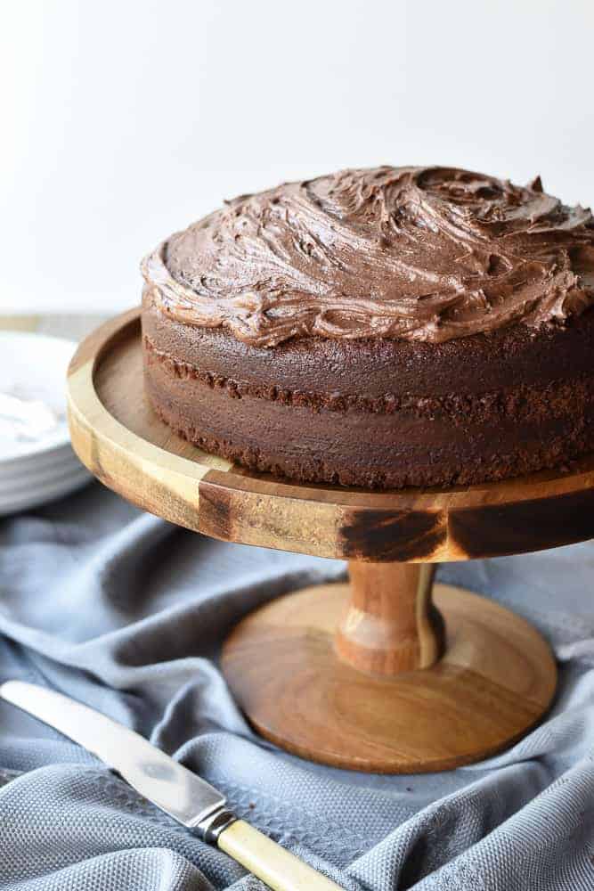 This homemade Chocolate Cake recipe is just as easy as a box cake but much more delicious! Topped with simple chocolate frosting, everyone will be asking for seconds! #homemade #chocolatecake #homemadecake #homemaderecipe #cakerecipe Frosted Chocolate cake on wooden cake stand