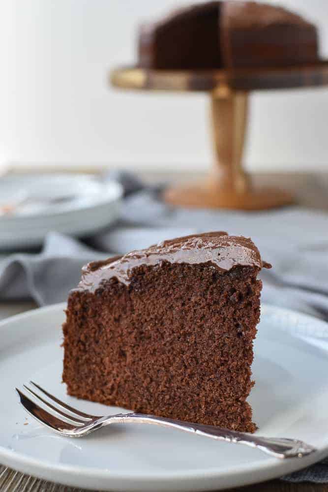 This homemade Chocolate Cake recipe is just as easy as a box cake but much more delicious! Topped with simple chocolate frosting, everyone will be asking for seconds! #homemade #chocolatecake #homemadecake #homemaderecipe #cakerecipe Slice of chocolate cake on white plate with cake fork, whole cake in the background.