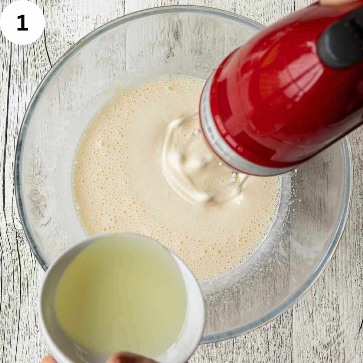 Adding oil to a bowl of frothy, creamy mixture using an electric mixer.