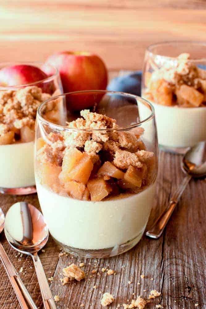 Cinnamon Panna Cotta with Apple Crumble topping makes a perfectly easy dessert that is as impressive as it is flavourful! #pannacotta #cinnamon #pannacottarecipe #italiandessert #applecrumble #applecrisp Three glasses of panna cotta topped with caramelised apples and crumble; two red apples in the background