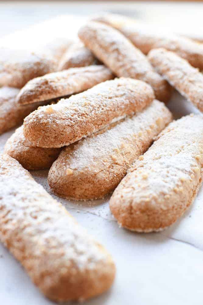 Sponge fingers are light, airy and actually quite easy to make at home! You might know this Italian treat by their other name - savoiardi or even lady fingers, either way you'll love the homemade taste of this recipe. #spongefingers #ladyfingers #savoiardi #biscotti #savoiardibiscuits #italianrecipe #italianfood #baking #desserts pile of savoiardi dusted with powdered viewed from an angle.