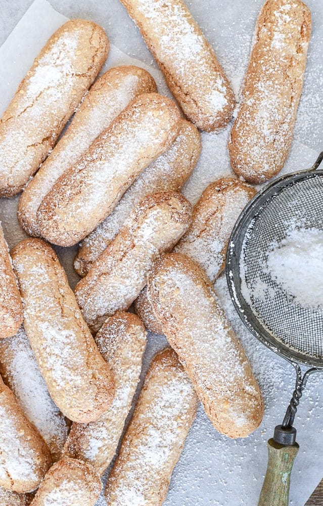 Sponge fingers are light, airy and actually quite easy to make at home! You might know this Italian treat by their other name - savoiardi or even lady fingers, either way you'll love the homemade taste of this recipe. #spongefingers #ladyfingers #savoiardi #biscotti #savoiardibiscuits #italianrecipe #italianfood #baking #desserts pile of oblong cookies dusted with powdered sugar and sieve on the side viewed from above.