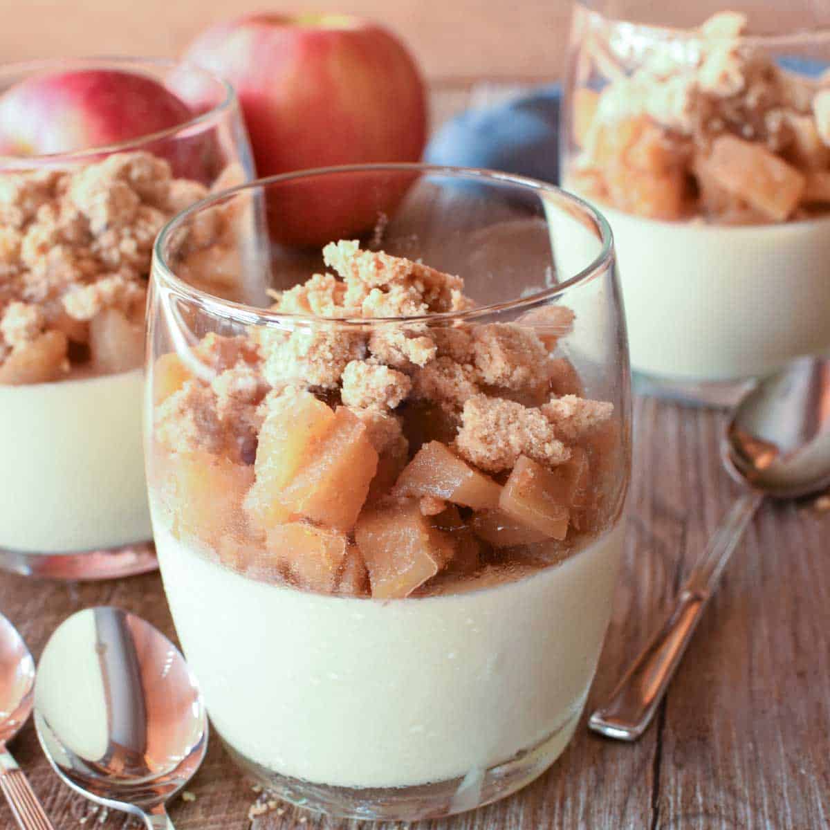 Three glasses of panna cotta topped with caramelised apples and crumble; two red apples in the background.