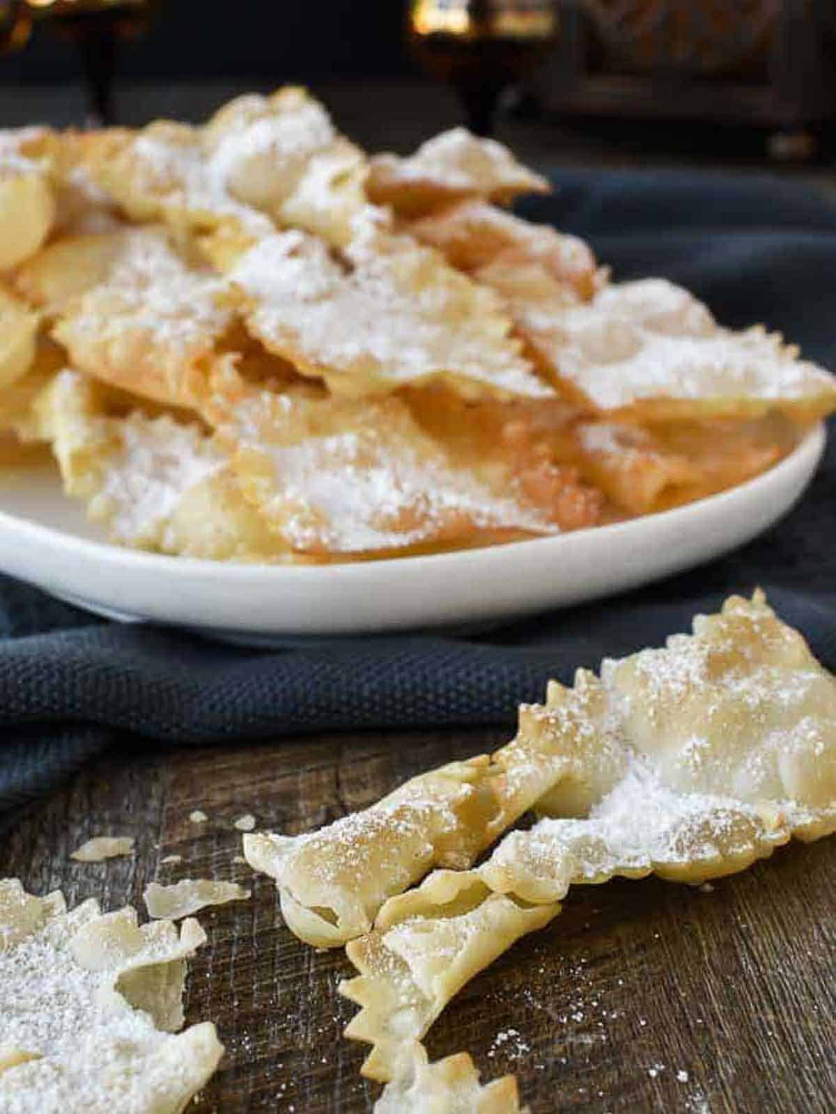 Chiacchiere di Carnevale are a crispy fried Italian pastry which are popular during carnvale and many other celebrations. Try stopping at just one - impossible! #crostoli #angelwings #Italianfritters #easter #christmasrecipes #lent #Italiancookie #italianrecipes Plate of fried pastry cookies dusted with powdered sugar.