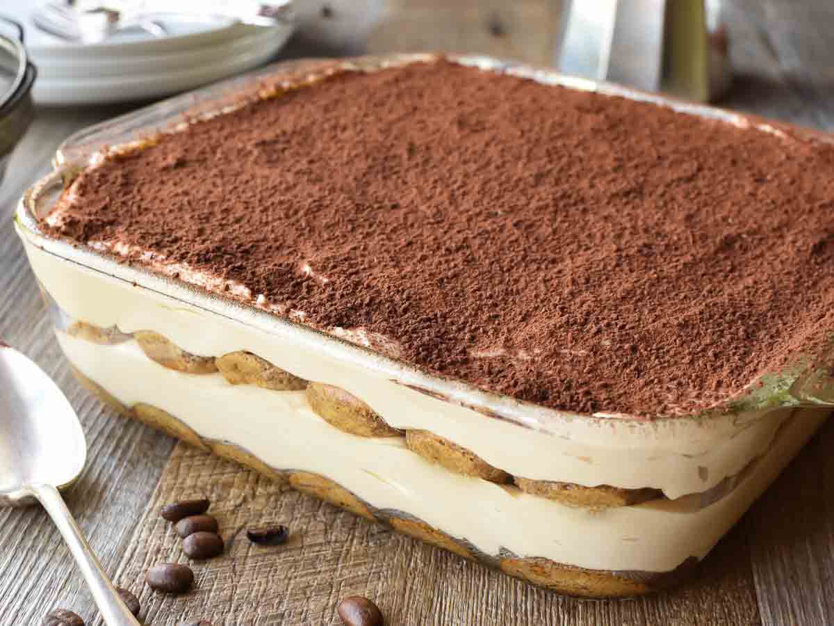 Tiramisu is a much loved Italian dessert. But do you know how to make tiramisu at home? Follow my recipe for delicious results! #tiramisu #dessert #italian #cream #nobake #tiramisurecipe #italiandessert Cocoa topped, creamy dessert in square glass pan viewed from the side.