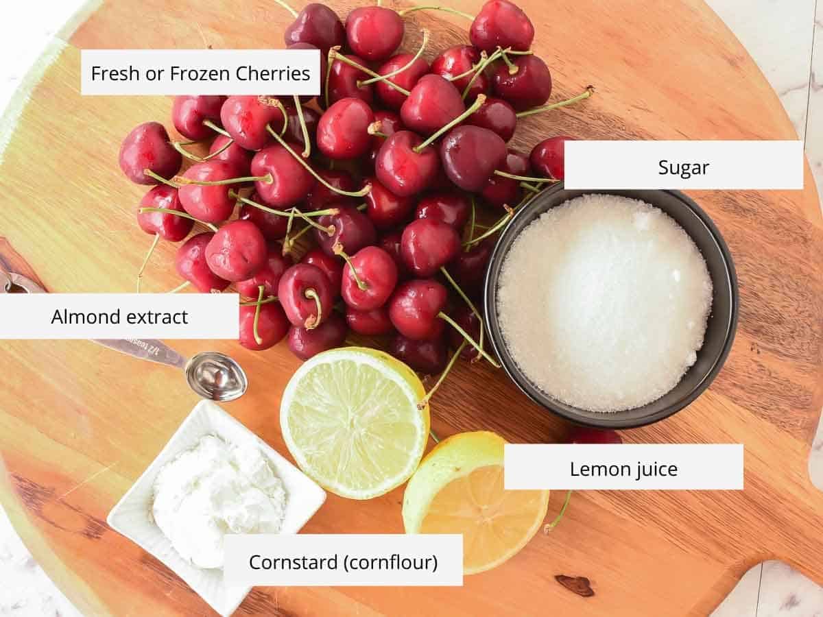Cherry Sauce using only 5 ingredients is easy to make at home. Perfect to use for cakes, cheesecakes, ice cream, pancakes, waffles or any dessert. #cherrysauce #easy #recipe #fresh#frozen #icecream #cheesecake #sweet #best #cherry #fruitsauce #dessert Fresh cherries, sugar, cut lemon, metal spoon and cornstarch on wooden board viewed from above.
