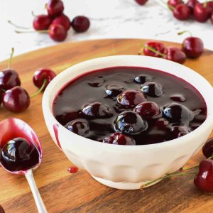 Cherry sauce in white bowl with cherries scattered around, spoonful of sauce beside bowl