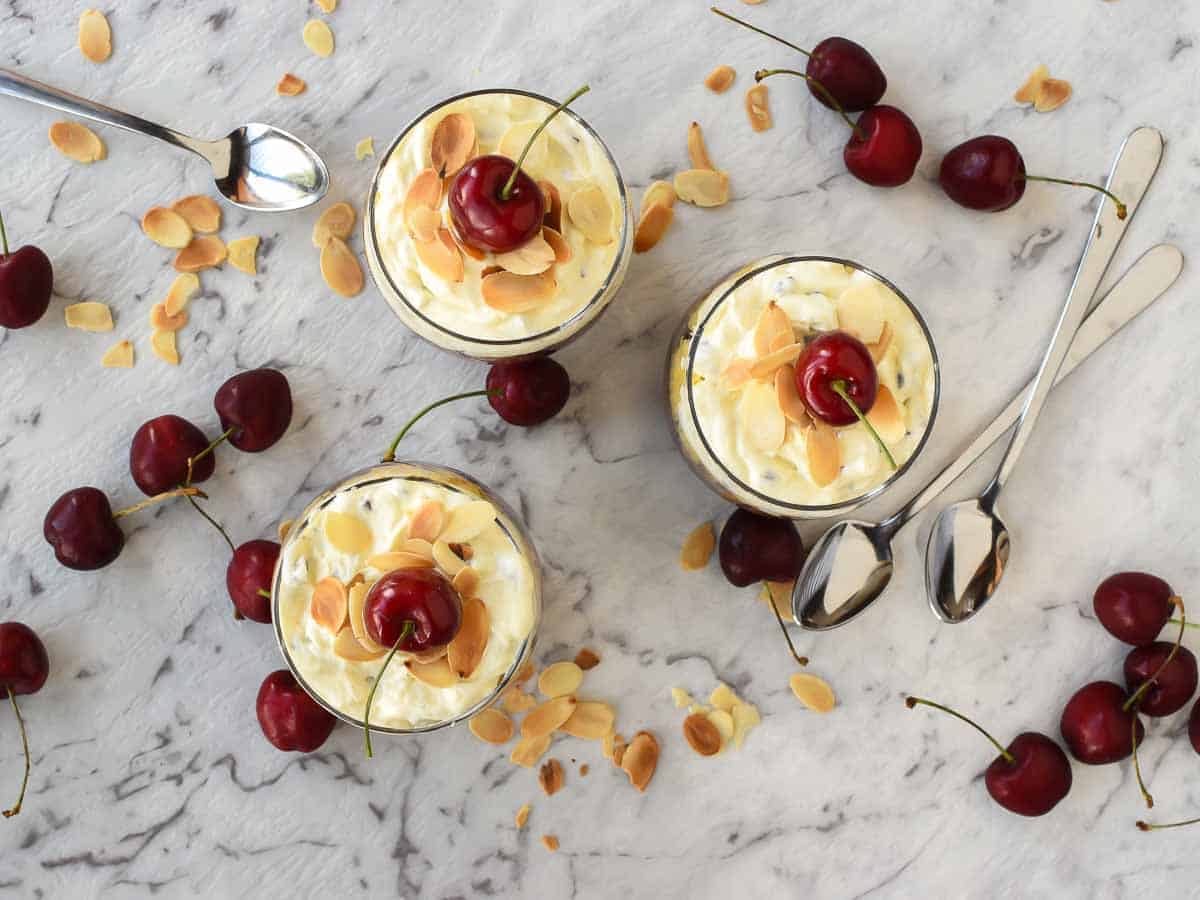 Trifle Desserts with Mascarpone and Cherries! These individual Mascarpone and cherry trifles are so delicious! Using store bought cake, pandoro or brioche these are filled with creamy mascarpone and cherry sauce. Perfect for that special trifle dessert recipe! #trifle #christmastrifle #dessertcups #christmasdessert #cherrysauce #fruitsauce #easy #recipe #christmas Three glasses of creamy trifle dessert with cherries scattered around viewed from above.