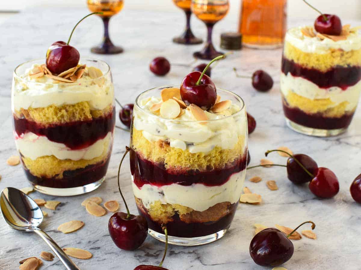 Trifle Desserts with Mascarpone and Cherries! These individual Mascarpone and cherry trifles are so delicious! Using store bought cake, pandoro or brioche these are filled with creamy mascarpone and cherry sauce. Perfect for that special trifle dessert recipe! #trifle #christmastrifle #dessertcups #christmasdessert #cherrysauce #fruitsauce #easy #recipe #christmas Layered creamy cherry dessert in a glass with a cherry on top cherries scattered around and two other glasses of dessert in the background.