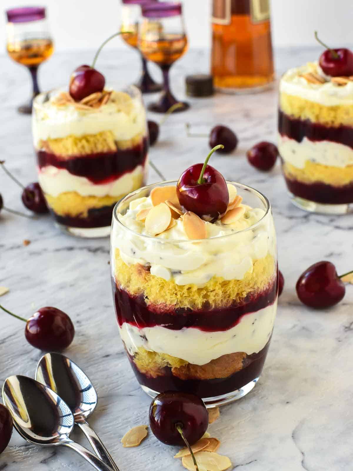 Trifle Desserts with Mascarpone and Cherries! These individual Mascarpone and cherry trifles are so delicious! Using store bought cake, pandoro or brioche these are filled with creamy mascarpone and cherry sauce. Perfect for that special trifle dessert recipe! #trifle #christmastrifle #dessertcups #christmasdessert #cherrysauce #fruitsauce #easy #recipe #christmas Close up of mini trifles layered in a glass with a cherry on top, cherries scattered around and two other glasses of dessert in the background.