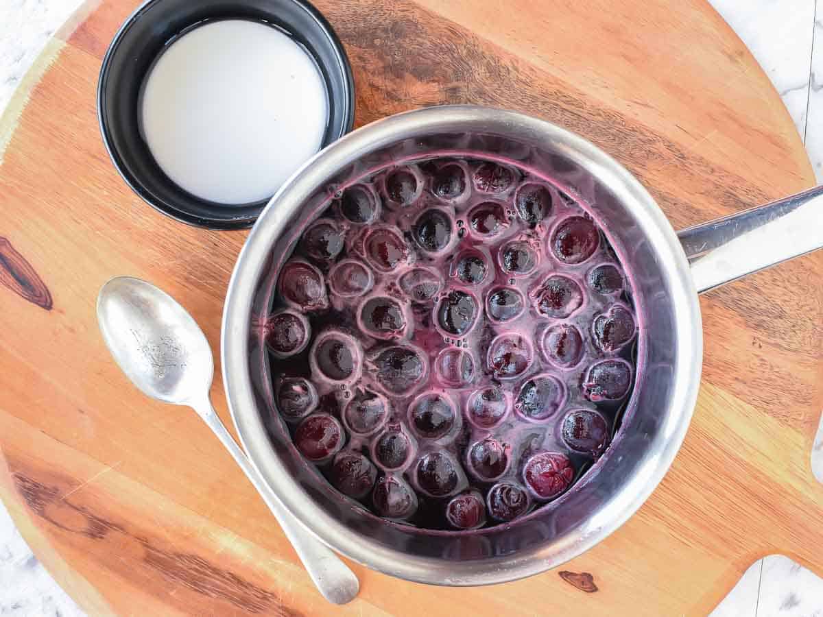 Cherry Sauce using only 5 ingredients is easy to make at home. Perfect to use for cakes, cheesecakes, ice cream, pancakes, waffles or any dessert. #cherrysauce #easy #recipe #fresh#frozen #icecream #cheesecake #sweet #best #cherry #fruitsauce #dessert Cherries and liquid in a pot with white cornstarch slurry in black bowl and metal spoon beside viewed from above.