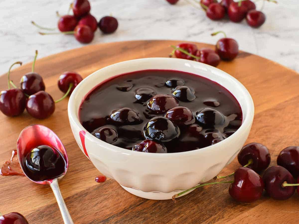 Cherry Sauce using only 5 ingredients is easy to make at home. Perfect to use for cakes, cheesecakes, ice cream, pancakes, waffles or any dessert. #cherrysauce #easy #recipe #fresh#frozen #icecream #cheesecake #sweet #best #cherry #fruitsauce #dessert Cherry sauce in white bowl with cherries scattered around, spoonful of sauce beside bowl.