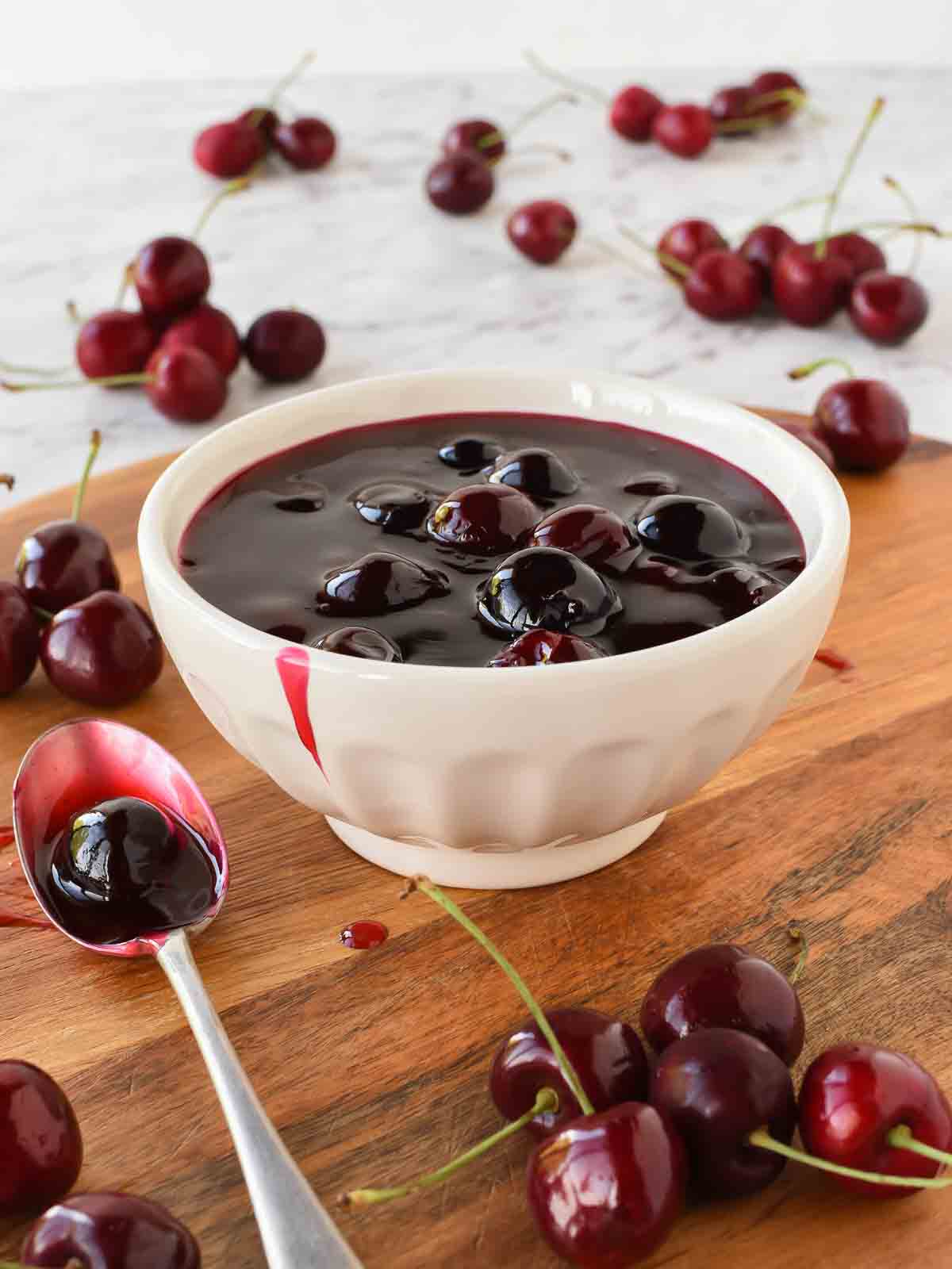 Cherry Sauce using only 5 ingredients is easy to make at home. Perfect to use for cakes, cheesecakes, ice cream, pancakes, waffles or any dessert. #cherrysauce #easy #recipe #fresh#frozen #icecream #cheesecake #sweet #best #cherry #fruitsauce #dessert Cherry sauce in white bowl with cherries scattered around, spoonful of sauce beside bowl.