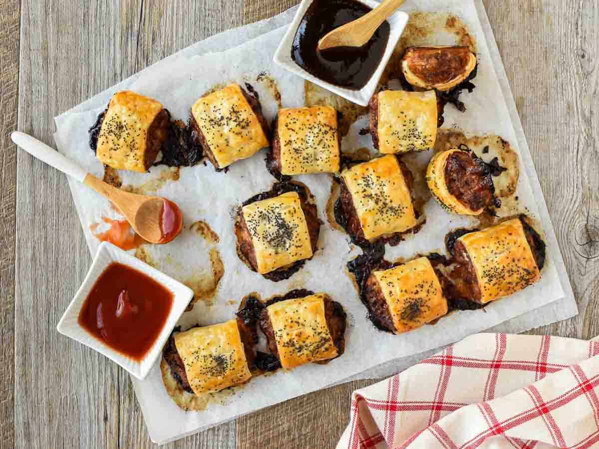 A Sausage Roll recipe with cheese and bacon that is so full of flavour like you've never tasted before. Plus this recipe is not that difficult to make! #sausagerolls #recipe #partyfood #entertaining #homemade sausage rolls on baking paper with black sauce and red sauce viewed from above