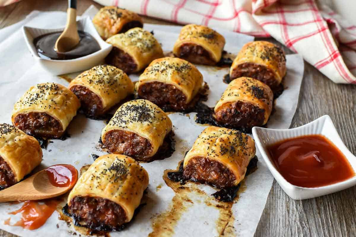 A Sausage Roll recipe with cheese and bacon that is so full of flavour like you've never tasted before. Plus this recipe is not that difficult to make! #sausagerolls #recipe #partyfood #entertaining #homemade sausage rolls on baking paper with black sauce and red sauce
