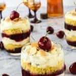 layered creamy cherry dessert in a glass with a cherry on top cherries scattered around and two other glasses of dessert in the background