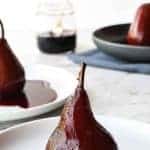 red wine poached pone red wine poached pear sitting upright in red syrup on a white plate with another plated pear in the background and two more pears in a grey bowl to the side