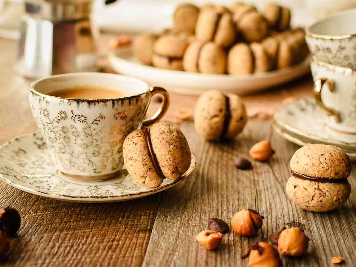 Hazelnut Cookie Sandwich with chocolate on the edge of a cup and saucer with more in the background.