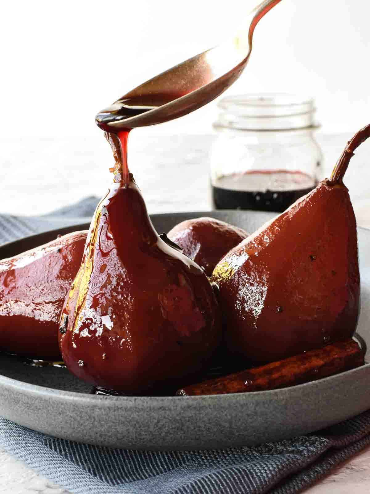 Poached Pears in Red Wine and Spices is a simple and impressive dessert that is equally at home at a dinner party or family meal. These delicious pears glisten with syrup and are fragrant with spices. Included is a special ingredient that balances out the sweetness. #pears #pearrecipe #redwine #dessert #recipe #poachedpears 4 poached pears in red wine in a grey shallow bowl with spoon pouring red syrup over a pear