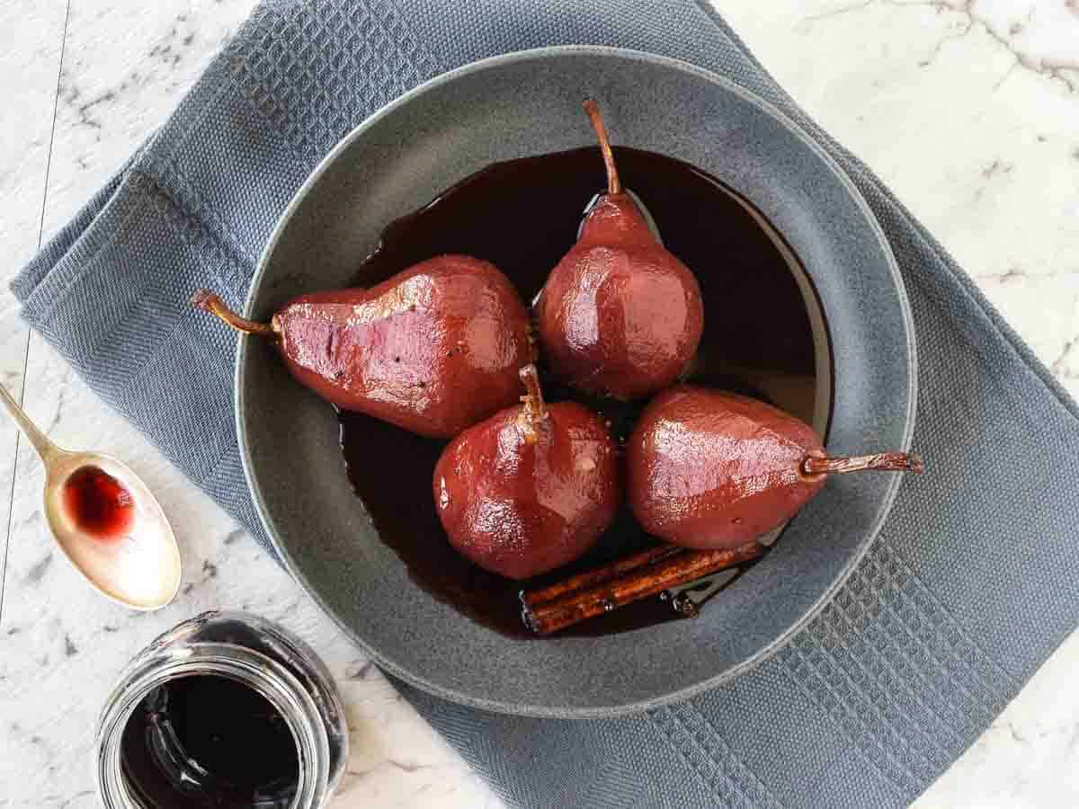 Poached Pears in Red Wine and Spices is a simple and impressive dessert that is equally at home at a dinner party or family meal. These delicious pears glisten with syrup and are fragrant with spices. Included is a special ingredient that balances out the sweetness. #pears #pearrecipe #redwine #dessert #recipe #poachedpears Four finished pears in a grey bowl viewed from above